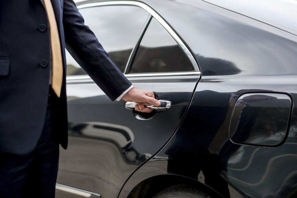 A chauffeur in a suit holding the rear door handle of a black sedan.